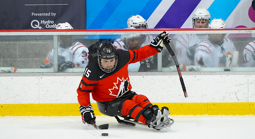Un joueur de parahockey de l'équipe canadienne manie la rondelle devant le banc des joueurs des États-Unis.