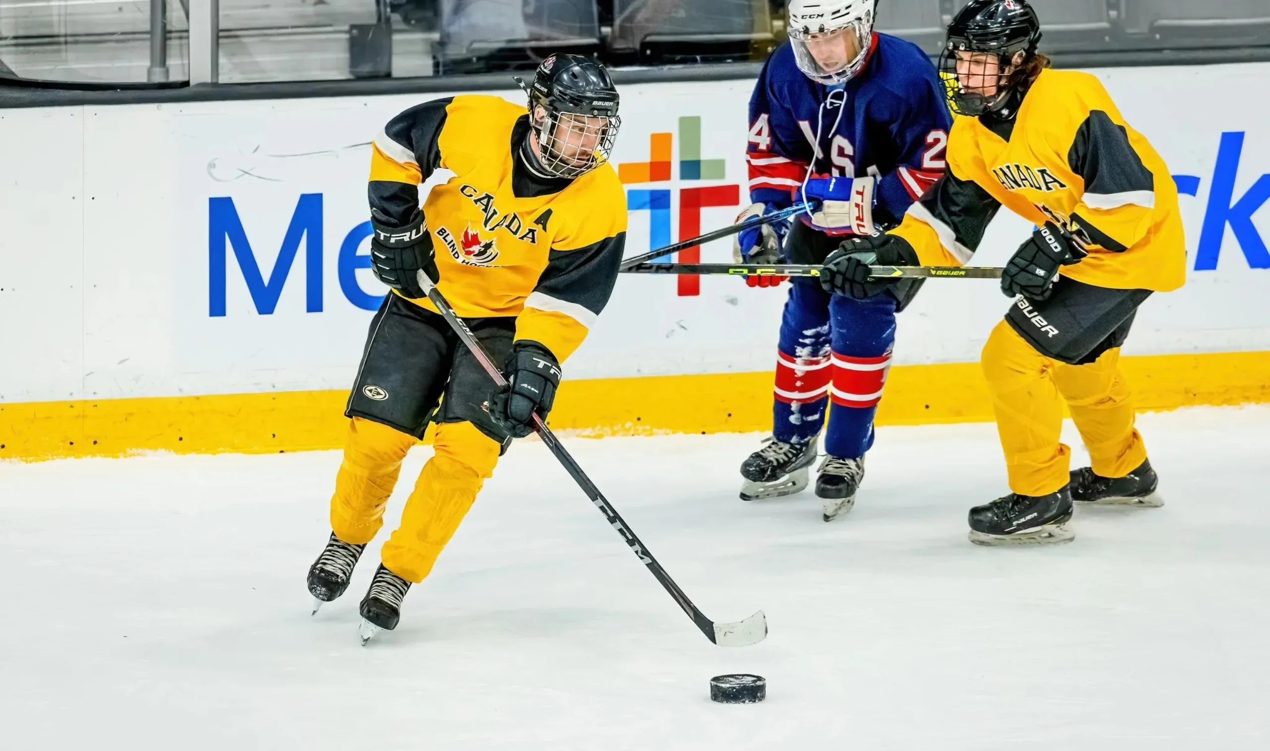 Un joueur de l'Équipe canadienne de hockey sonore vêtu de jaune patine avec la rondelle, poursuivi par un joueur de l'équipe adverse vêtu de bleu et défendu par une joueuse de son équipe.