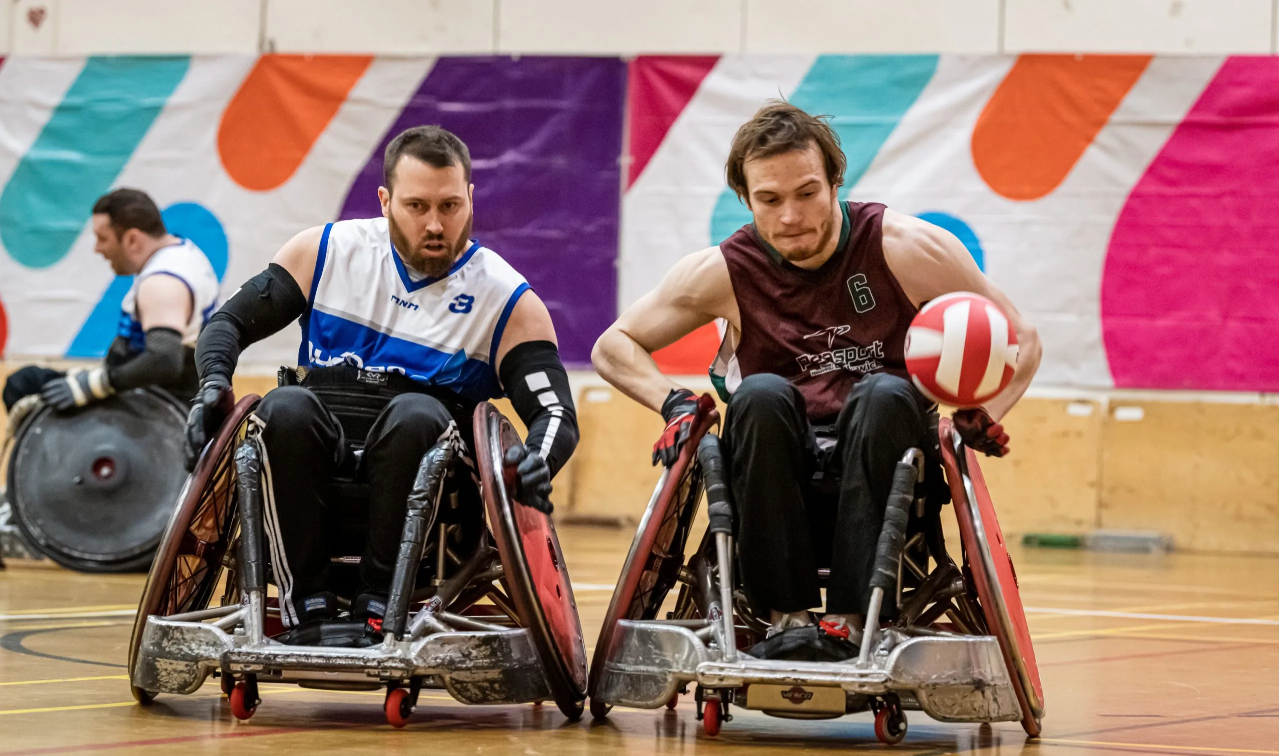 Deux athlètes de rugby en fauteuil roulant se déplacent vers la caméra. Celui de droite tient le ballon, tandis que celui de gauche essaie de lui enlever.