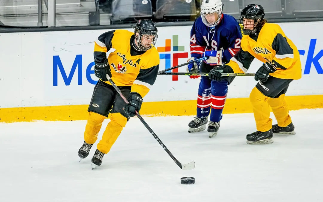 Un joueur de l'Équipe canadienne de hockey sonore vêtu de jaune patine avec la rondelle, poursuivi par un joueur de l'équipe adverse vêtu de bleu et défendu par une joueuse de son équipe.