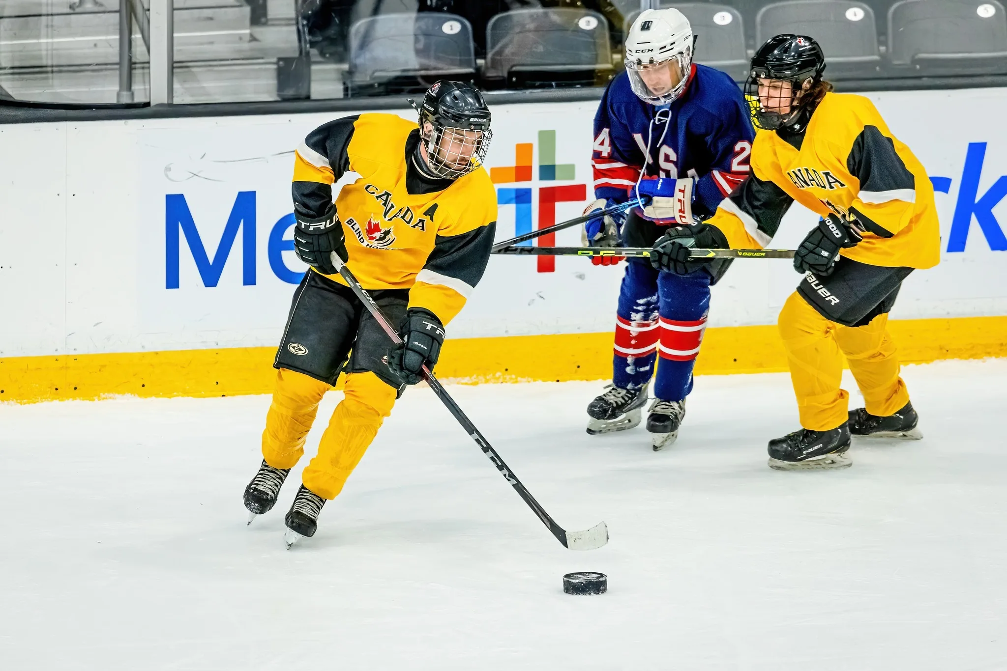 Un joueur de l'Équipe canadienne de hockey sonore vêtu de jaune patine avec la rondelle, poursuivi par un joueur de l'équipe adverse vêtu de bleu et défendu par une joueuse de son équipe.