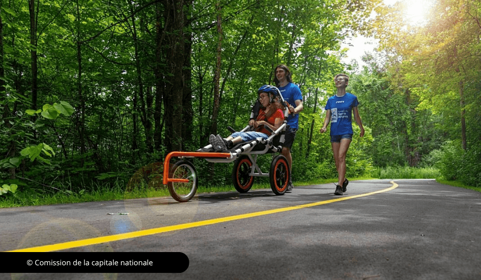 Deux jeunes marchent avec une femme wui utilise un fauteuil de randonnée sur un sentier asphalté.